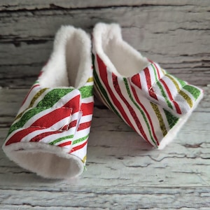 以下が含まれることがあります： 赤、緑、金色のストライプ模様のベビーブーティーのペア。内側は白いフェイクファーで裏打ちされています。ブーティーは柔らかく柔軟なソールで設計されています。