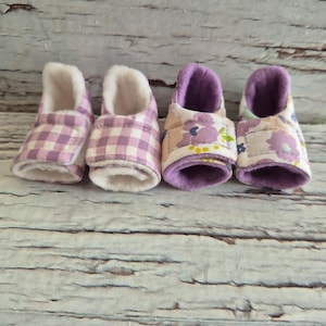 以下が含まれることがあります： 4つのベビーブーティー。2つは薄紫と白のギンガムチェック、2つは紫色の内装のフローラル柄です。すべて白くふわふわの裏地が付いています。明るい木目調の背景に配置されています。