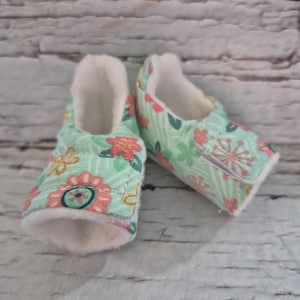 以下が含まれることがあります： 緑、ピンク、黄色を基調とした花柄のベビーブーティーのペア。ブーティーは柔らかい白い内側とベルクロ留めです。背景は明るい色の木製の表面です。