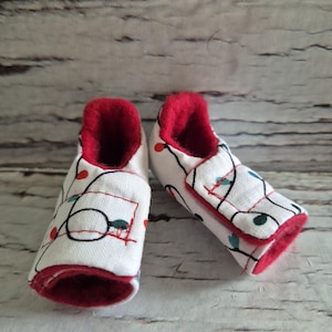 以下が含まれることがあります： 白地に赤色の内装のベビーブーティーのペア。ブーティーは、黒いラインのデザインと小さな赤とターコイズのアクセントが特徴です。ブーティーは柔らかい生地で作られています。