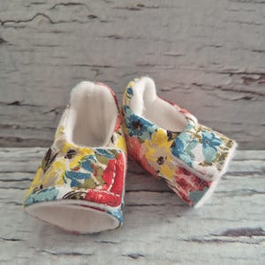 May include: A pair of baby shoes with a floral pattern in red, yellow, blue, and white. The shoes have a white interior and a soft, padded sole. The shoes are designed for infants.