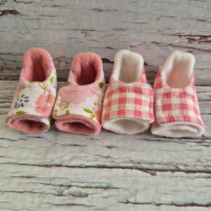 May include: Four baby booties in a row, each with a different pink pattern. The first two have floral designs, the third has a pink and white gingham pattern, and the fourth has a smaller pink and white pattern. All have a soft, white interior.