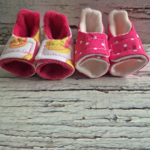 以下が含まれることがあります： ベビーブーティー4足。2足は白地に黄色とオレンジのデザイン。他の2足は、白い水玉模様の明るいピンク色です。すべて柔らかい白い内側とピンクのトリムが付いています。