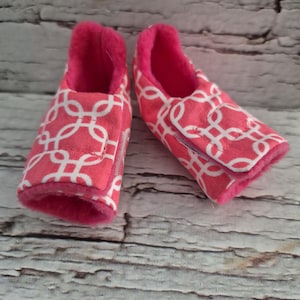 以下が含まれることがあります： 白の幾何学模様が入ったピンク色のベビー用スリッパのペア。スリッパは柔らかいピンク色の内側と面ファスナー留めが付いています。柔らかい模様の生地で作られています。