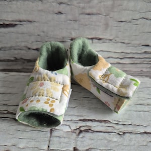 以下が含まれることがあります： 白地に黄色、桃色、緑色の花柄のベビーブーティーのペア。内側は無地グリーンで、面ファスナーで留めます。背景は明るい色の風化した木材です。