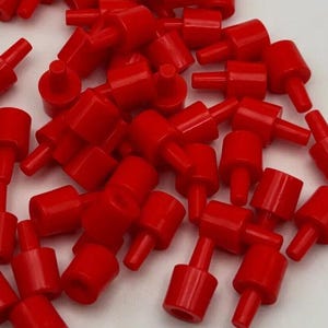 以下が含まれることがあります： 鮮やかな赤色の小さなプラスチック製ペグの集まりで、それぞれ円筒形のベースと突き出たステムが特徴です。これらの均一な部品は山積みになっており、光沢のある仕上げと、さまざまな組み立て用途に適した一貫した形状を示しています。