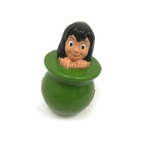 Puede incluir: Una figura de plástico verde de un personaje con cabello negro y una cara sonriente, emergiendo de una olla verde. La figura tiene un diseño de dibujos animados y está sobre un fondo blanco.