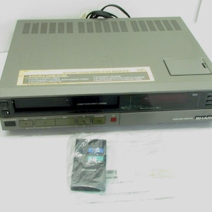 May include: A grey Sharp VHS video cassette recorder with a wireless remote control. The VCR has a slot for inserting tapes, a digital display, and several control buttons. The remote is in a plastic bag.