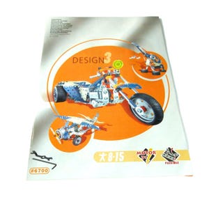 May include: A sheet with instructions for a metal construction set. The sheet features illustrations of a motorcycle, airplane, and robot. The word "DESIGN 3" is in an orange circle. The age range is 8-15. The set includes a wrench and the number 6700.