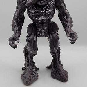 Peut inclure: Une figurine en plastique violet foncé représentant une créature au torse musclé et texturé, aux épaules larges et aux mains griffues. La figurine présente une structure de tête large et distincte avec une ouverture centrale et des jambes épaisses.