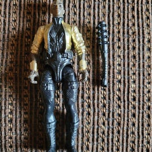Peut inclure: Une figurine en plastique avec une coiffure mohawk, portant une veste dorée métallique sur une combinaison noire texturée avec des bottes et des gantelets noirs assortis. La figurine est accompagnée d'un accessoire massue noire à pointes.