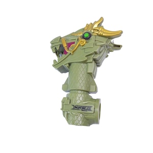 Puede incluir: Un juguete verde con forma de dragón con detalles dorados y un ojo verde. El juguete tiene un aspecto texturizado y escamoso, y la palabra "NINJAGO" está impresa en la base. La boca del dragón está abierta, revelando un interior rosa.