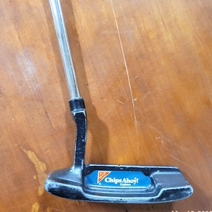 Op de afbeelding: Een zwarte metalen golfputter met een zilverkleurige schacht. De kop van de putter is voorzien van een blauw rechthoekig inzetstuk met het oranje en witte Chips Ahoy! Cookies-logo. De kop van de club vertoont gebruikssporen met zichtbare verfschade.