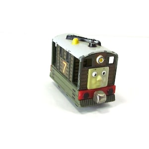 May include: A toy train carriage with a cartoon face. The carriage is predominantly green and brown, with a grey roof and a red front. The number 7 is displayed on the side. A yellow detail is on the roof.