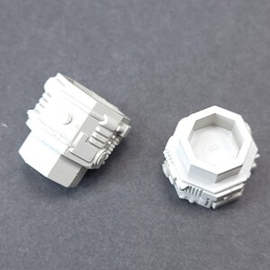 May include: Two small, light grey, geometric-shaped components. One is a rectangular prism with a cylindrical top and detailed side panels. The other is a hexagonal shape with a detailed base. Both are on a grey surface.