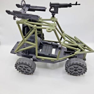Peut inclure: Un véhicule militaire miniature doté d'un châssis noir et d'une cage de protection vert olive. Le véhicule est équipé de deux fusils jouets noirs montés sur le cadre supérieur et d'un volant placé entre eux. Il possède quatre roues noires.