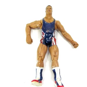 Puede incluir: Una figura de acción de plástico de un hombre musculoso con un maillot y botas de lucha libre azules y rojas. La figura tiene articulaciones y posa con los brazos extendidos. El maillot tiene estrellas blancas.