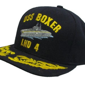 May include: A navy blue baseball cap featuring yellow embroidered text that reads USS BOXER and LHD 4. An embroidered graphic of a gray naval ship is positioned between the text. The flat brim displays yellow embroidered leaf-like detailing.