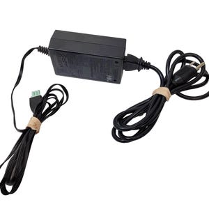 Op de afbeelding: Een zwarte voedingsadapter met een rechthoekige behuizing en bevestigde zwarte snoeren. Eén snoer heeft een groene connector en de andere een standaard stekker. De snoeren zijn gebundeld met bruine bandjes.