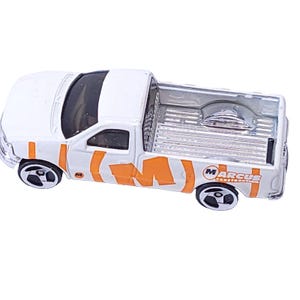 Può includere: Un camioncino giocattolo bianco con dettagli arancioni e ruote nere. Il camion ha la scritta "Marcus Construct" sul lato. È una replica in miniatura di un veicolo reale.