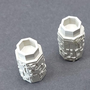 May include: Two light grey, cylindrical objects with a geometric design. Each has a hexagonal top and a detailed, textured body. The objects are set against a plain, grey background.