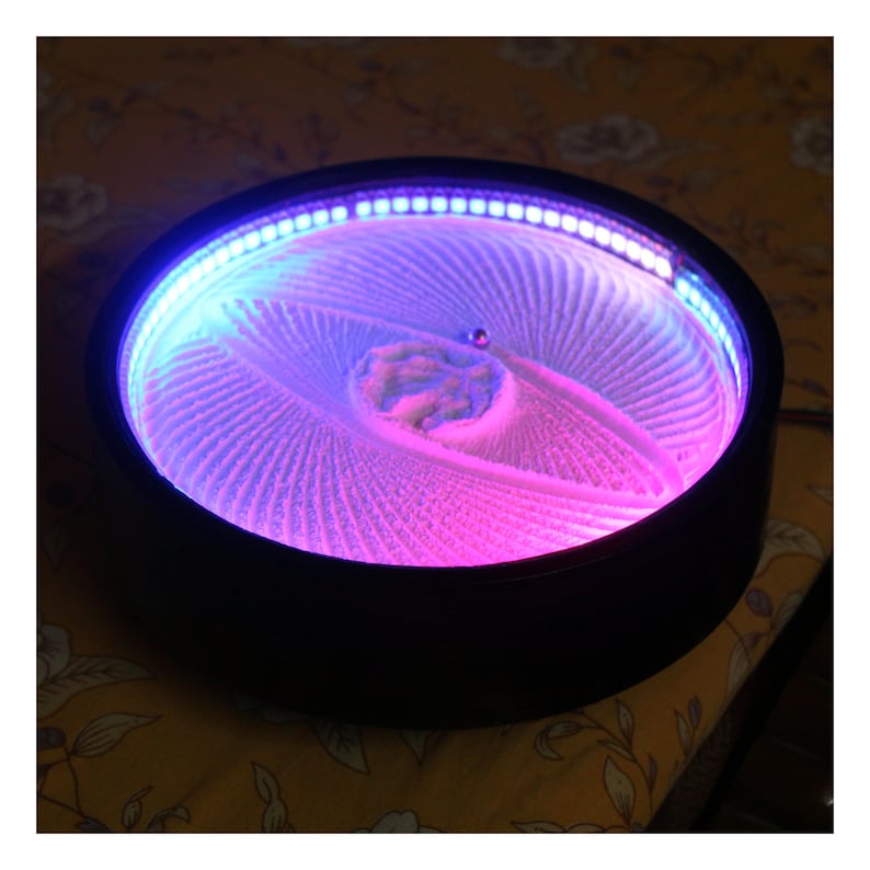 K&ouml;nnte beinhalten: Eine schwarze, kreisf&ouml;rmige kinetische Sandkunst-Anzeige mit einem faszinierenden Muster. Der Sand wird von einem Farbverlauf aus rosa und blauen LED-Leuchten am Rand beleuchtet. Eine kleine Kugel rollt &uuml;ber den Sand und erzeugt komplizierte Designs.