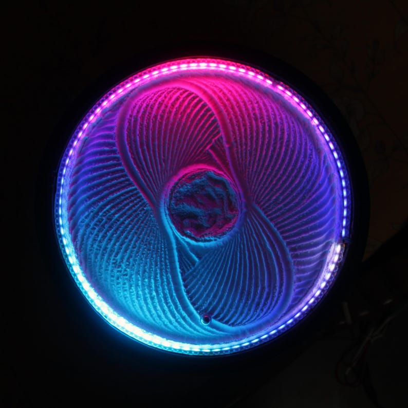 K&ouml;nnte beinhalten: Runde, beleuchtete Sandkunst-Anzeige mit einem faszinierenden Muster. Der Sand ist in wirbelnden Linien geformt, beleuchtet von einem lebendigen Farbverlauf aus rosa und blauen LED-Leuchten. Der Hintergrund ist dunkel.