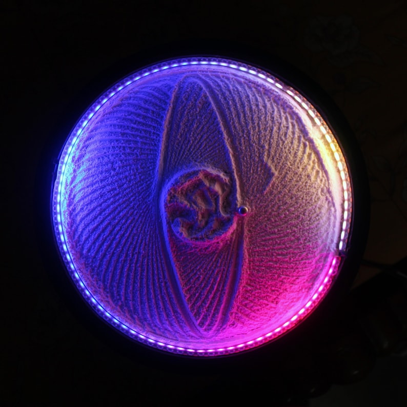 K&ouml;nnte beinhalten: Ein kreisf&ouml;rmiges kinetisches Sandkunstwerk mit schwarzem Rahmen und leuchtendem LED-Rand. Die Sandkunst zeigt ein Spiralmuster mit blauer und rosa Beleuchtung, wodurch ein faszinierender visueller Effekt entsteht. Das Kunstwerk ist vor einem dunklen Hintergrund platziert.