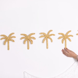 May include: Six woven palm tree wall decorations in a light tan colour are arranged horizontally on a white wall. A hand is reaching towards the rightmost tree. The trees are made of a woven material.