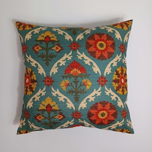 May include: Square decorative pillow with a teal background and a repeating floral pattern. The pattern includes orange, yellow, and brown flowers, with cream-colored scrollwork accents. The pillow has a textured appearance.