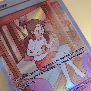 Op de afbeelding: Een cartoonillustratie van een meisje met bruin haar en een wit shirt dat een kaart vasthoudt waarop staat: "Shiny Arc: Schiet een magische pijl die door het hele veld van je tegenstander gaat en elk Pok&eacute;mon 100 schade toebrengt."