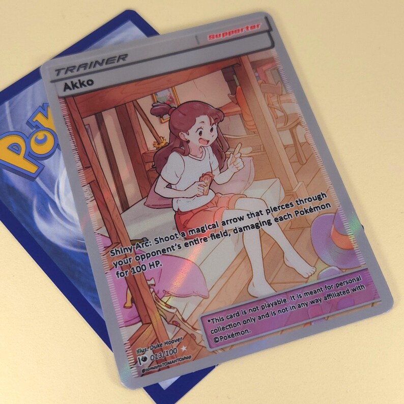 Op de afbeelding: Een Pokemon trading card met een cartoonillustratie van een meisje met bruin haar en een roze jurk. De kaart heeft de titel "Akko" en de tekst "Shiny Arc: Schiet een magische pijl die door het hele veld van je tegenstander gaat en elk Pokemon 100 HP schade toebrengt." De kaart is een supporterkaart.