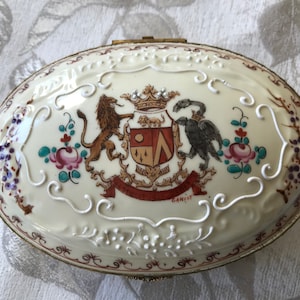 Könnte beinhalten: Eine ovale Porzellan-Schmuckdose mit weißem Hintergrund und goldener Einfassung. Der Deckel zeigt ein handgemaltes Wappen mit einem Löwen und einem Vogel, umgeben von floralen Motiven und weißen Schnörkeln. Die Inschrift "GANIST" ist am unteren Rand des Wappens sichtbar.