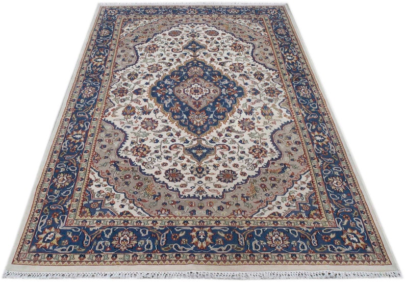 Puede incluir: Una alfombra de color crema con un patr&oacute;n floral azul y rojo. La alfombra tiene un medall&oacute;n central con un dise&ntilde;o azul y blanco. La alfombra tiene un borde de flecos.
