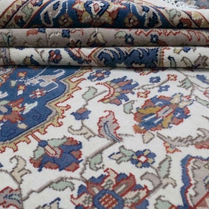 Puede incluir: Una alfombra blanca con un patr&oacute;n floral azul, rojo y dorado. La alfombra est&aacute; doblada en tres partes, mostrando los intrincados detalles del dise&ntilde;o.