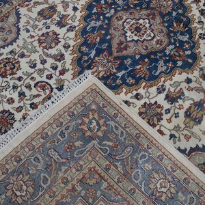 Puede incluir: Una alfombra de color crema con un patr&oacute;n floral azul y rojo. La alfombra tiene un borde de flecos.