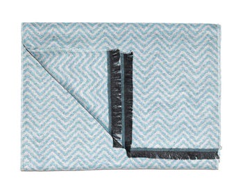 May include: A folded, rectangular throw blanket with a chevron pattern in light blue and white. The blanket has a dark gray border and fringe along one edge. The blanket is made of a soft, woven material.