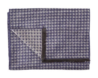 May include: A folded, rectangular blanket with a houndstooth pattern in shades of blue and beige. The blanket has a dark fringe along one edge. The fabric appears soft and warm, suitable for use as a throw or decorative piece.
