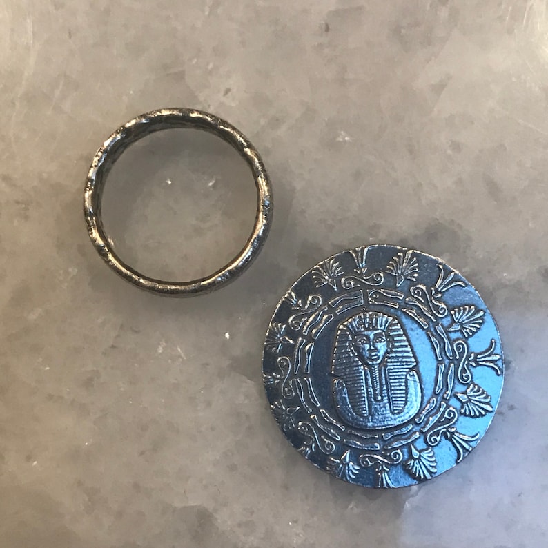 May include: A silver ring with a textured surface and a silver coin with an embossed image of a pharaoh's head surrounded by a decorative border.