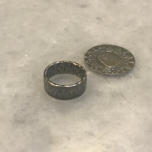 May include: A silver ring made from a coin with an intricate design. The ring is sitting on a white marble surface next to a silver coin.