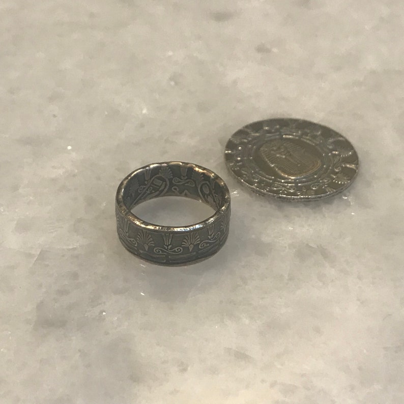 May include: A silver ring made from a coin with an intricate design. The ring is sitting on a white marble surface next to a silver coin.