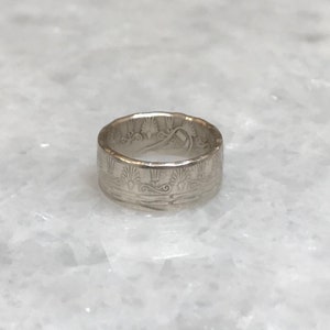 May include: A silver ring with a detailed design of a stylized wave pattern and a central motif of a tree or plant.