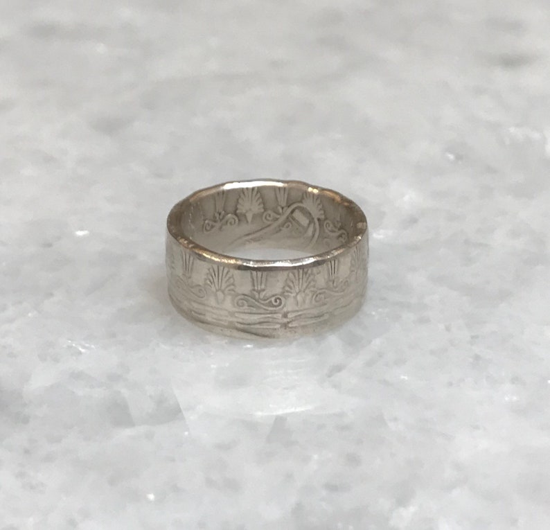 May include: A silver ring with a detailed design of a stylized wave pattern and a central motif of a tree or plant.