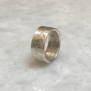 May include: A silver ring with an intricate design around the band. The design features a repeating pattern of stylized flowers and leaves.