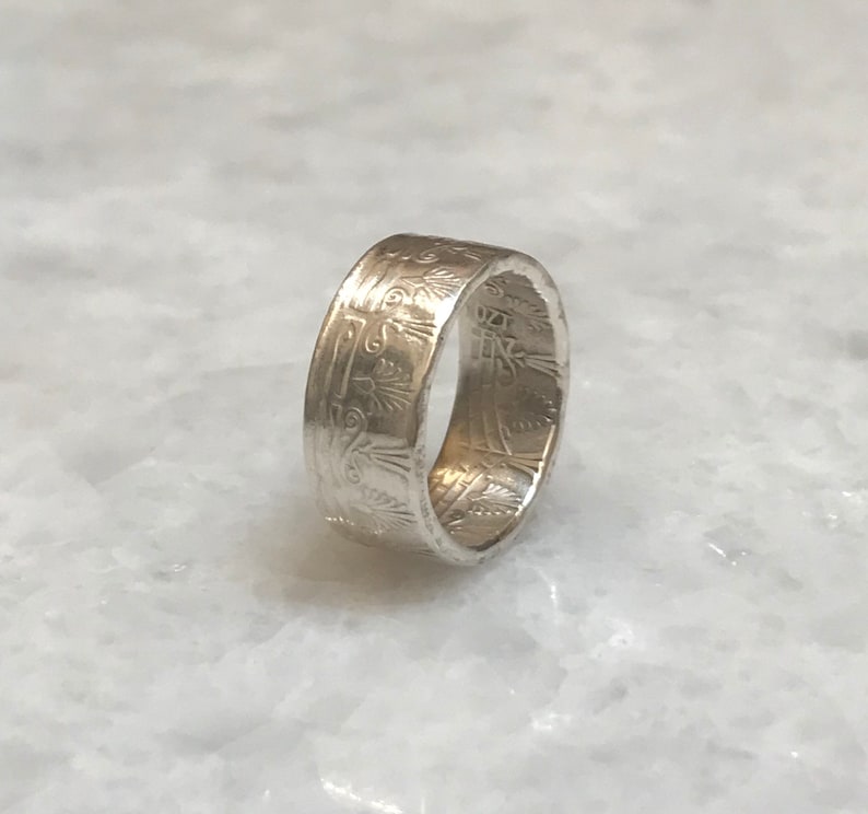 May include: A silver ring with an intricate design around the band. The design features a repeating pattern of stylized flowers and leaves.