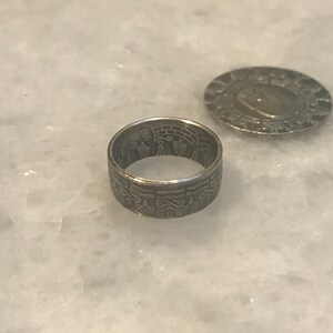 May include: A silver ring made from a coin with an intricate design featuring a tree and other symbols.