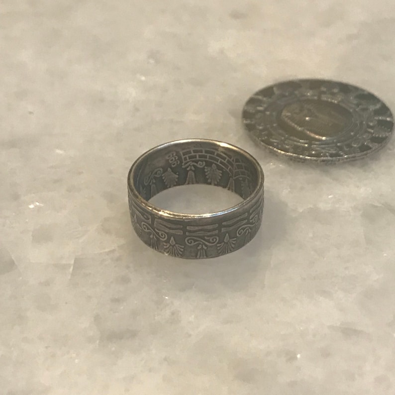 May include: A silver ring made from a coin with an intricate design featuring a tree and other symbols.