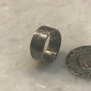 May include: A silver ring made from a coin with an intricate design of an Egyptian symbol. The ring is on a white surface.