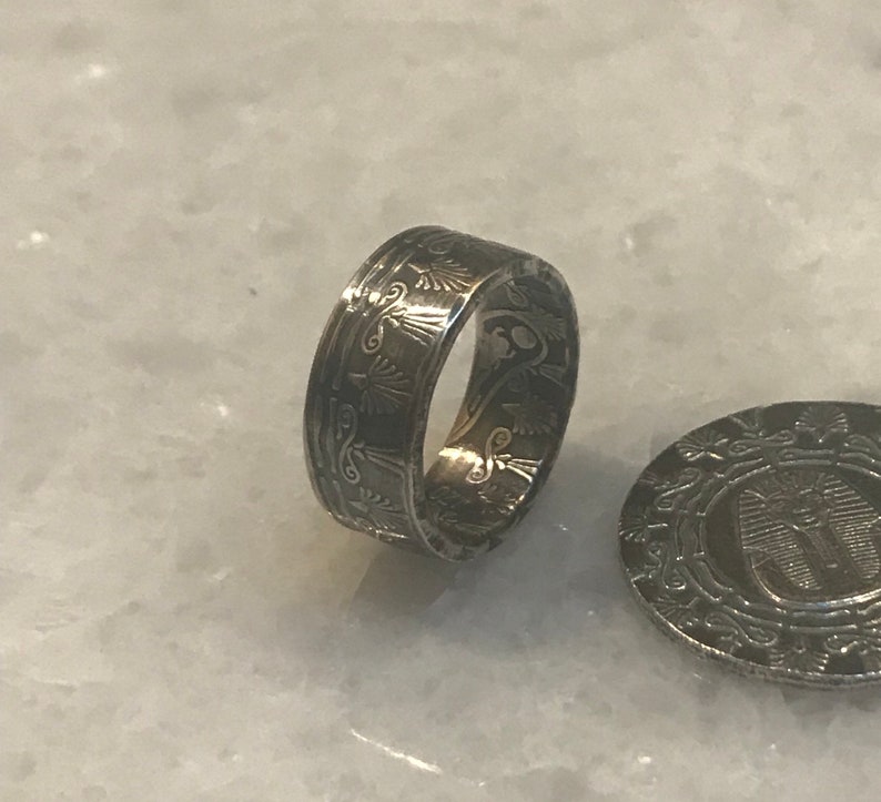 May include: A silver ring made from a coin with an intricate design of an Egyptian symbol. The ring is on a white surface.