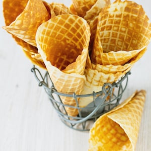 May include: A group of six empty waffle cones in a wire basket. The cones are made of a light brown waffle material and are arranged in a pile.
